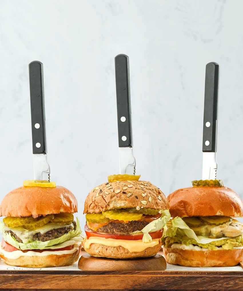 Three gourmet burgers with pickles and steak knives on a wooden board at Pawdy Neighbors.