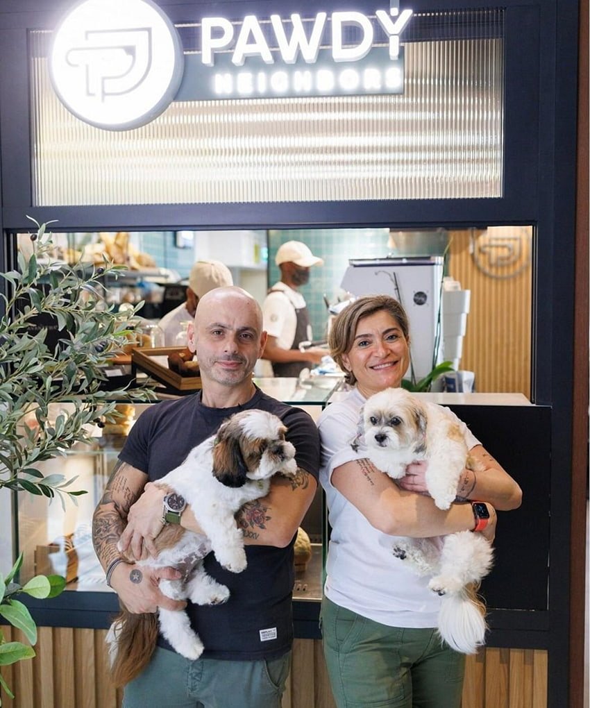 A couple smiling and holding two small fluffy dogs outside a pet-friendly café.