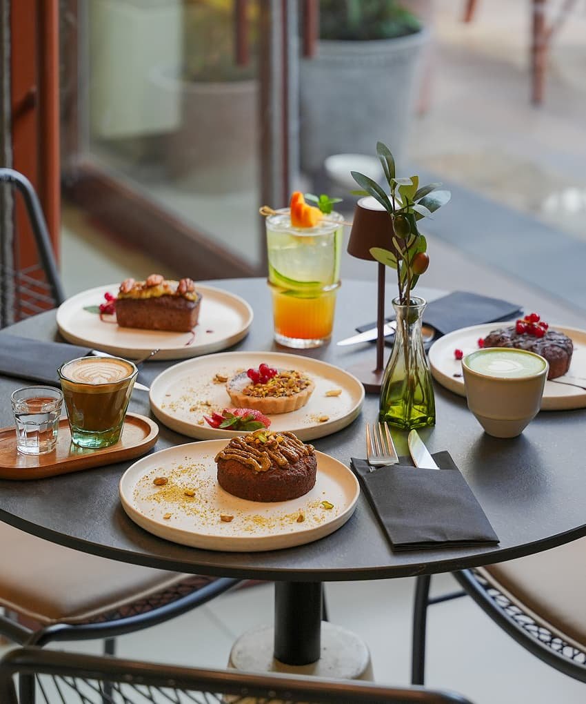 Overhead view of a café table spread with pastries, desserts, lattes, and a fruity cocktail on ceramic plates.