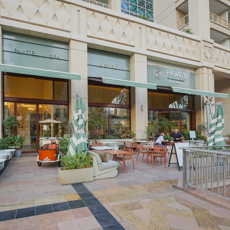Exterior of Pawdy Neighbors bistro and Pawtel Spa with outdoor patio seating, green-striped umbrellas, and a vintage orange food cart