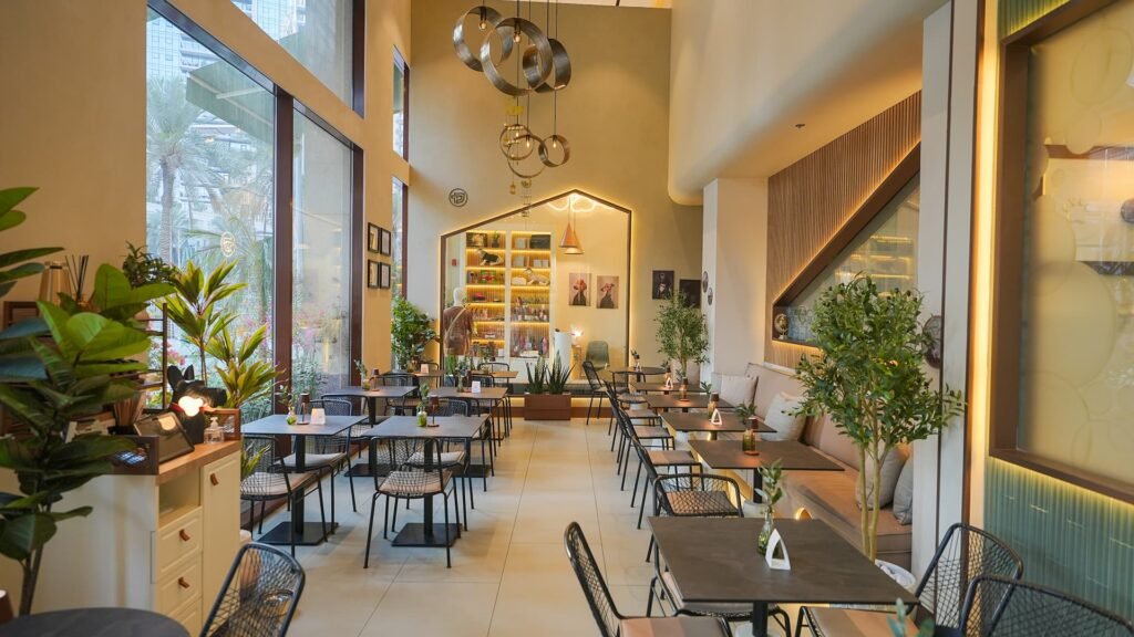 Spacious restaurant interior with tall ceilings, indoor plants, dark dining tables, and modern pendant lighting.